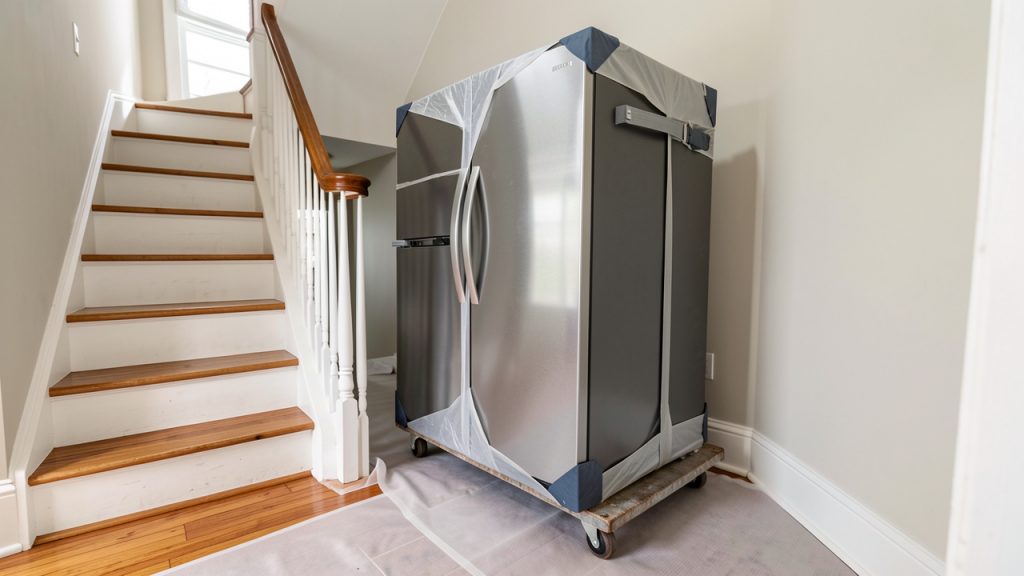 Un grand réfrigérateur protégé par des couvertures et installé sur un diable au pied d’un escalier, avec une protection couvrant le sol.